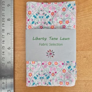 May include: A folded fabric selection with a floral pattern in pink, yellow, and teal on a white background. The fabric is wrapped with a grey band that reads "Liberty Tana Lawn Fabric Selection."