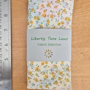 May include: A folded piece of Liberty Tana Lawn fabric with a floral print in shades of yellow, pink, and blue. The fabric is labeled "Liberty Tana Lawn Fabric Selection".