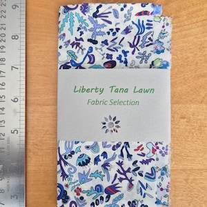 May include: A folded fabric selection with a floral pattern in shades of blue, purple, and teal on a white background. The fabric is wrapped with a gray band that reads "Liberty Tana Lawn Fabric Selection."