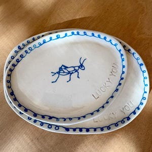 May include: Two white oval ceramic plates with a blue insect illustration and the words "LUCKY YOU". The plates have a blue dotted border.