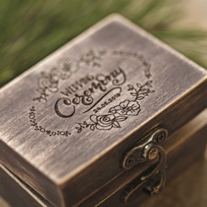 Peut inclure: Une boîte à alliances en bois avec un couvercle à charnière et un loquet en laiton. Le dessus de la boîte est gravé des mots "Our Wedding Ceremony" et de la date "24.05.2019" entourés d'un motif floral.