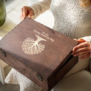 May include: A rectangular dark brown wooden box with a hinged lid and metal latches. The top of the box features a laser-engraved white tree design with the text The Kissane Family and EST. 2022 printed in a clean, sans-serif font.