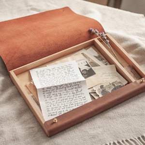 Peut inclure: Boîte en bois ouverte avec un couvercle en cuir marron, remplie de photographies anciennes et de lettres manuscrites. La boîte a un cadre en bois clair et une poignée en cuir marron foncé. Une brindille de lavande séchée repose à l'intérieur de la boîte, sur une couverture.