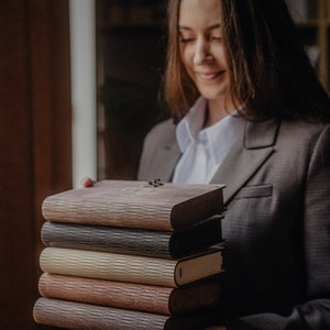 May include: A stack of six brown, black, and beige journals with a wooden cover. The journals are bound with a metal clasp and have a textured surface.
