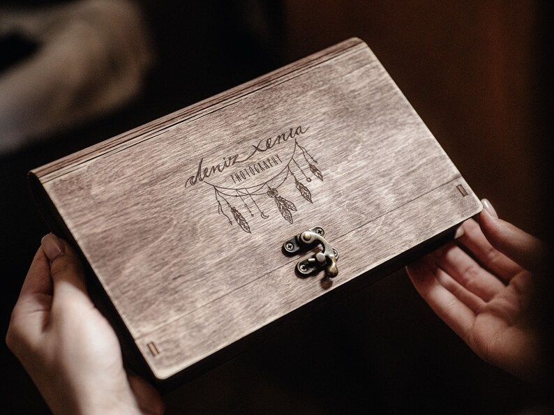 May include: A wooden box with a latch closure. The box is engraved with the text "deniz xenia photography" and a design of a dreamcatcher with feathers.