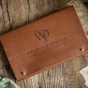 May include: A brown leather wedding album with the text "Wedding Pixels - Best Moments of the Day" and gold-colored rivets. The album is lying on a wooden surface with decorative elements.