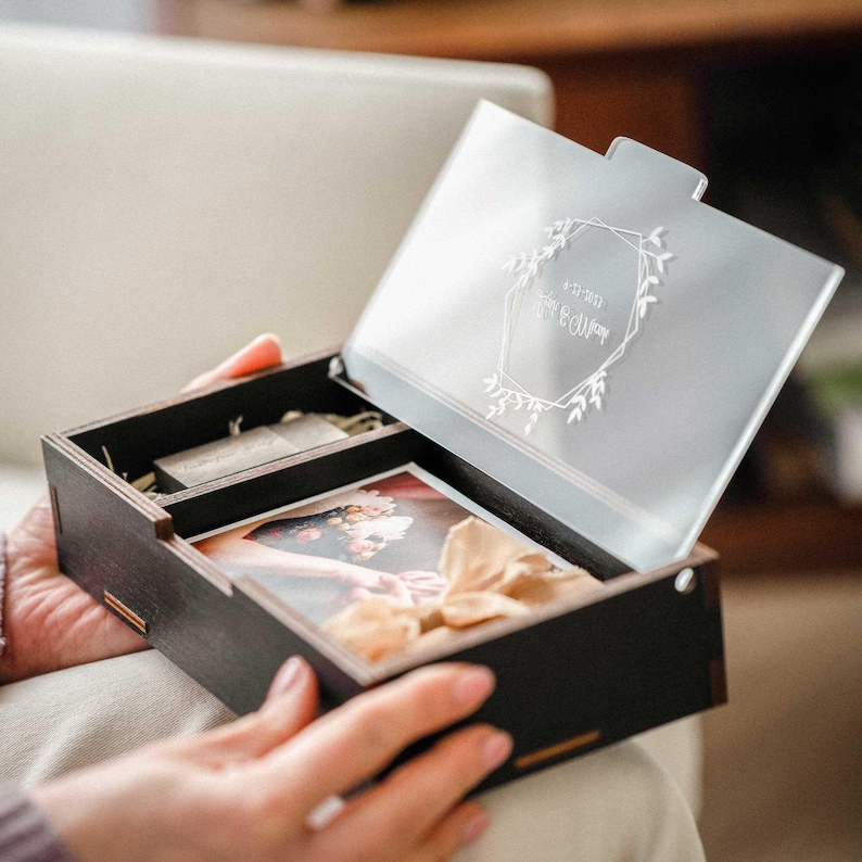Hands opening black wooden photo box with acrylic lid, showing 4x6 photo stack and USB compartment.