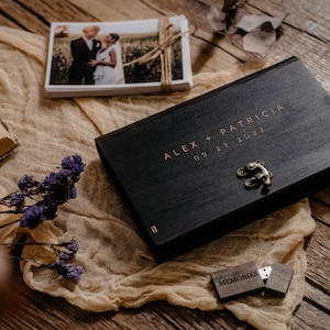 May include: A black wooden box with a gold latch and the inscription "ALEX + PATRICIA 09.23.2022". The box is sitting on a beige fabric surface with a small wooden USB drive labeled "MEMORIES" next to it.