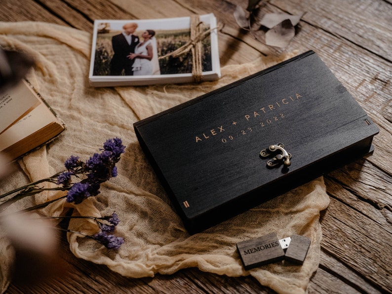 May include: A black wooden box with a gold latch and the inscription "ALEX + PATRICIA 09.23.2022". The box is sitting on a beige fabric surface with a small wooden USB drive labeled "MEMORIES" next to it.