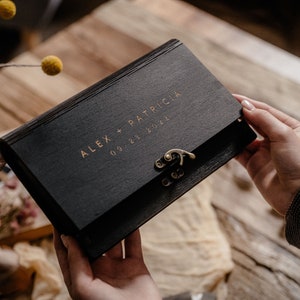 May include: A black wooden box with a gold latch and inscription "ALEX + PATRICIA 09.23.2022".