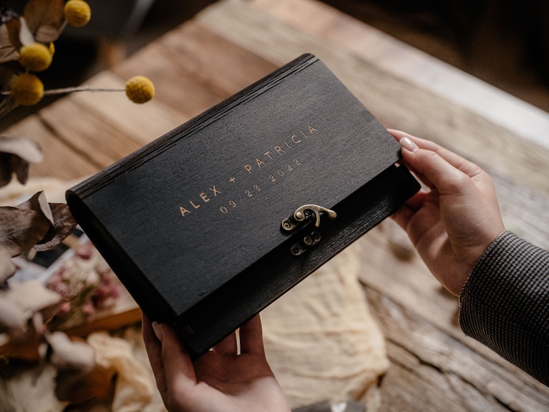 May include: A black wooden box with a gold latch and inscription "ALEX + PATRICIA 09.23.2022".