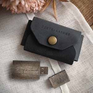 May include: A black leather USB drive case with the names "DEETS + KASSIEE" and the date "12-31-23" embossed on the front. A wooden USB drive with the names "Sherry + Jacob" engraved on it is next to the case.