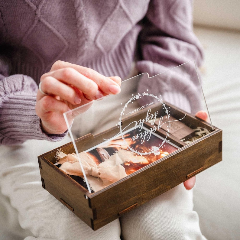 ands holding open walnut photo box with acrylic lid and 4x6 prints inside, ribbon and wood shavings visible.