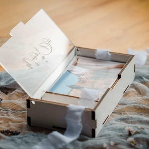 White wooden photo box open with clear engraved acrylic lid and 4x6 prints, ribbon styling.