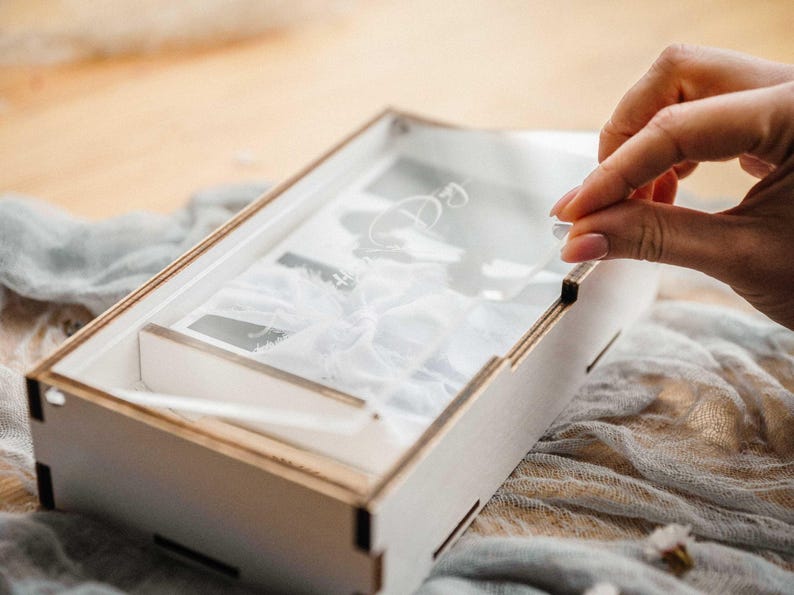 Hand lifting engraved acrylic lid on white photo box, showing photo storage and USB compartment.