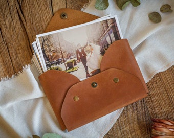 May include: A brown leather envelope with a flap closure and brass accents. The envelope is filled with photographs of a wedding couple.