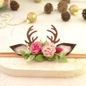 May include: A decorative headband featuring brown glitter antlers, brown ears, and pink and red flowers. The headband is set on a cream-colored base with a tan elastic band. Decorative pinecones and gold ornaments are in the background.