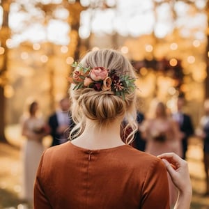 May include: A woman in a rust-colored dress with a floral hairpiece attends an outdoor wedding. The dress has long sleeves and a fitted waist. The hairpiece features pink and burgundy flowers and greenery. The background is blurred, with trees and other wedding guests visible.