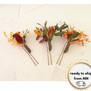 May include: Three floral hair pins with dried flowers in shades of orange, yellow, red, and green. The flowers are attached to brown stems and secured with copper-colored bobby pins. The hair accessories are arranged on a white surface.