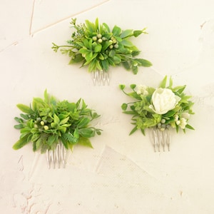 May include: Three floral hair combs with green leaves and white accents. One comb features a white flower. The combs have silver metal teeth. The hair accessories are arranged on a textured, off-white surface.