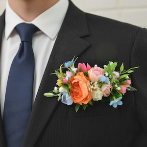 May include: A floral boutonniere featuring a large orange peony, smaller pink and cream roses, light blue blossoms, and green foliage. The arrangement is tucked into the breast pocket of a black pinstriped suit jacket worn with a white shirt and navy tie.