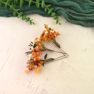 May include: Three hair pins with small bouquets of dried orange, white, and red flowers. The flowers are attached to brown bobby pins. Greenery and a dark green fabric are in the background.