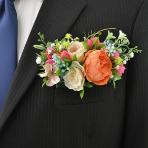 May include: A floral pocket square accessory featuring a cluster of artificial flowers in shades of orange, cream, pink, and light blue, accented with green foliage, tucked into the breast pocket of a black pinstriped suit jacket with a blue tie.