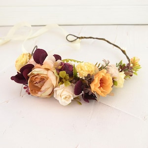 May include: A floral crown featuring an arrangement of artificial flowers in shades of deep burgundy, soft peach, cream, and yellow, accented with small green leaves and a thin brown wire base with long, light-colored ribbon ties at the ends.
