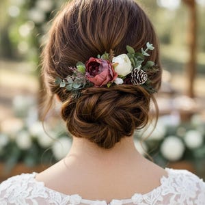 May include: A woman's brown hair is styled in a low bun, adorned with a floral hairpiece. The hairpiece features a burgundy peony, white flower, pinecone, and greenery. The woman wears a white lace dress.