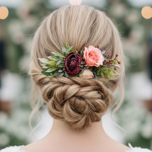 May include: A hair accessory featuring a cluster of artificial flowers including a deep burgundy rose, a soft peach rose, and various green leaves. The floral arrangement is secured into a braided blonde updo hairstyle with loose strands.