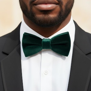 May include: A dark emerald green velvet bow tie worn with a crisp white button-down shirt and a black suit jacket. The bow tie features a smooth, soft-textured fabric with a classic pre-tied shape, centered perfectly at the collar of the shirt.