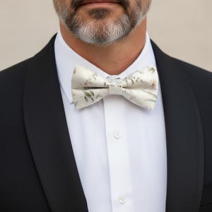 May include: A cream-colored bow tie with a floral pattern of pink and green leaves is the focal point of this image. The bow tie is worn with a white dress shirt and a black tuxedo jacket. The man in the image has a gray beard.