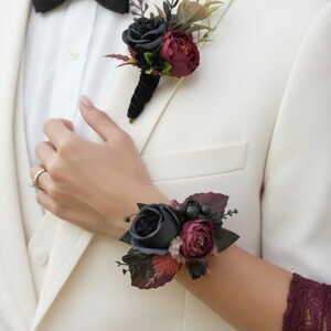 May include: A white suit with a black bow tie and floral accents. The boutonniere and wrist corsage feature dark blue and burgundy roses with greenery. The wrist corsage is worn on a burgundy lace sleeve.