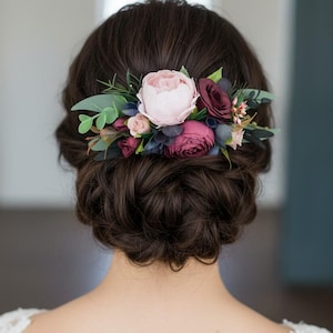 May include: A hair accessory featuring a cluster of artificial flowers including a large pale pink peony, deep burgundy roses, smaller pink buds, dark blue berries, and various green eucalyptus leaves arranged in a horizontal decorative spray.