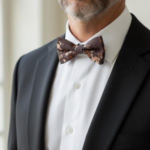 May include: A brown floral bow tie is the focal point of this image. The bow tie is paired with a white dress shirt and a black suit jacket. The floral pattern includes shades of brown and peach.