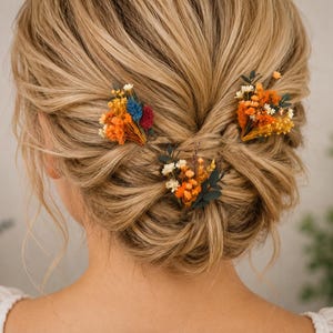 May include: Three hair pins featuring small bouquets of dried flowers in shades of orange, yellow, white, blue, and deep red, accented with dark green foliage. The floral arrangements are attached to pins designed to be secured into a hairstyle.