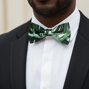 May include: A green and white leaf-patterned bow tie. The bow tie is worn with a white dress shirt and a black suit jacket. The bow tie's design features a tropical leaf pattern.