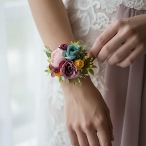 May include: A wrist corsage featuring a cluster of colorful artificial flowers. The arrangement includes pink, teal, burgundy, and yellow blooms, accented with green foliage. The corsage is secured on the wrist with a ribbon, perfect for weddings or special events.