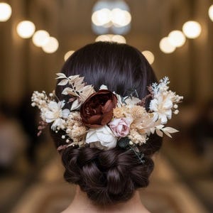 May include: A hair accessory featuring a collection of dried and silk flowers in shades of deep burgundy, soft pink, cream, and white. The arrangement is secured into a dark brown braided updo hairstyle, showcasing various textures of foliage and petals.