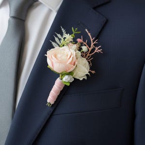 May include: A floral boutonniere featuring a pale pink rose, a small white rose, green foliage, and delicate pink twig accents. The stems are wrapped in a soft pink velvet ribbon, pinned to the lapel of a dark navy blue suit jacket.