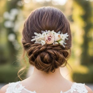 May include: A hair comb accessory featuring a cluster of soft pink and cream-colored artificial flowers, dried grasses, and white foliage. The decorative piece is secured into a brown hair bun, showcasing delicate petals and textured botanical elements.