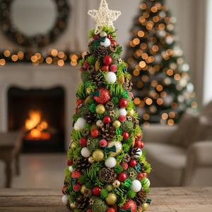 May include: A tabletop Christmas tree decoration featuring green pine needles, brown pinecones, and a mix of red, gold, and white spherical ornaments. The tree is topped with a woven white star and includes small green bows throughout the foliage.