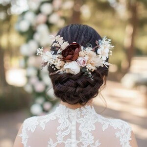 May include: A dark-haired person with a floral hairpiece, featuring a large burgundy flower and smaller white and pink blooms. The person wears a white lace dress with a high neckline and button detail down the back. The background is blurred.