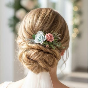 May include: A hair accessory featuring a light blue rose, a dusty pink rose, and green foliage tucked into a blonde updo hairstyle. The accessory is positioned above a white veil, with the hair styled in a low, intricate, twisted bun.