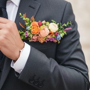 May include: A charcoal gray suit jacket with a white shirt and a dark tie, featuring a pocket square made of a cluster of artificial flowers in shades of orange, pink, cream, purple, and blue, accented with green foliage tucked into the breast pocket.