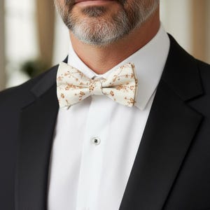 May include: A cream-colored bow tie with a delicate floral pattern in shades of brown. The bow tie is set against a crisp white dress shirt and a black tuxedo jacket, creating a formal and elegant look.