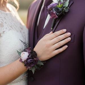 May include: A floral wrist corsage featuring dark purple and light pink roses with green foliage, paired with a matching boutonniere pinned to a deep purple suit jacket. The arrangement includes small dark berries and fern-like leaves.