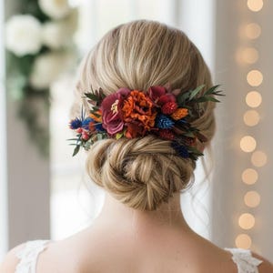 May include: A woman's blonde hair is styled in a low bun, adorned with a floral hairpiece. The hair accessory features burgundy, orange, and blue flowers with green foliage, creating a vibrant contrast against the light hair.