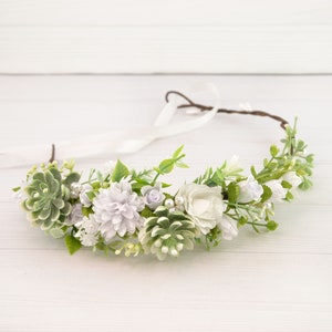 May include: A floral crown featuring a variety of white, light purple, and green artificial flowers and succulents. The crown is made with a brown wire base and white ribbon ties. The flowers include roses, small white blossoms, and succulent-like greenery.
