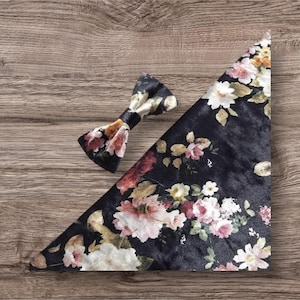 May include: A black floral bow tie and matching pocket square. The bow tie has a black band and the pocket square is triangular. The floral pattern includes pink, white, and yellow flowers on a black background. The items are displayed on a wooden surface.